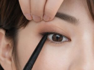 上まぶたを指で軽く持ち上げながら、アイライナーをまつ毛の生え際に当てている目元のアップ写真。まぶたを押さえて、アイラインを入れやすくする様子を示している